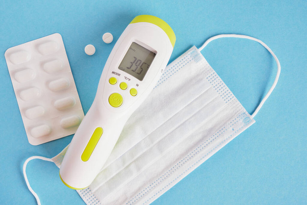pills, a face mask and an electronic infrared non-contact thermometer that shows a dangerously high temperature on a blue background, a body temperature of 39.5 degrees Celsius - Valokuva, kuva
