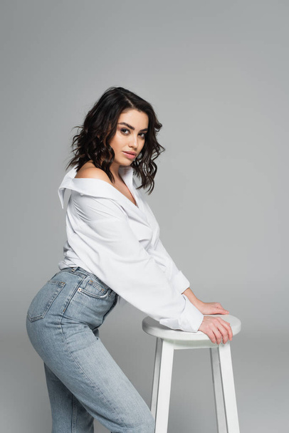 Young woman in white shirt looking at camera near chair isolated on grey  - Foto, Imagen