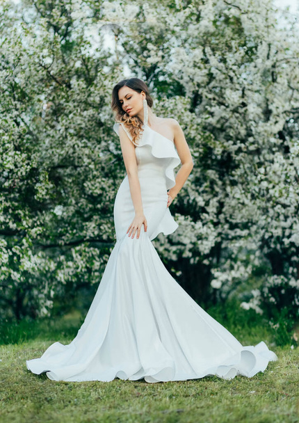 Young beautiful woman fashion model posing in white evening dress silhouette mermaid. Stylish girl bride perfect gown, long train. Nature background green blooming trees. Spring wedding cherry garden. - 写真・画像