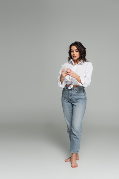 Pretty brunette woman in shirt and jeans using smartphone on grey background - Fotoğraf, Görsel