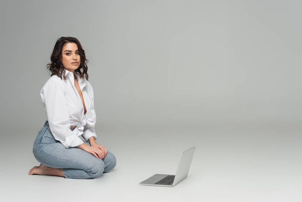 Verführerische Frau im weißen Hemd blickt in die Kamera neben dem Laptop auf grauem Hintergrund  - Foto, Bild