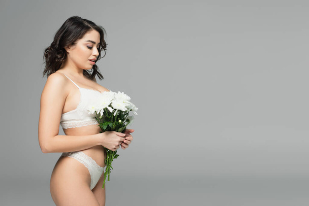 Side view of sensual woman in underwear holding white chrysanthemums isolated on grey - Foto, Imagen