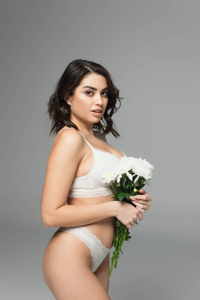 Sensual woman in lingerie holding bouquet and looking at camera isolated on grey - 写真・画像