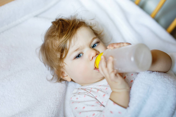 Χαριτωμένο κοριτσάκι κρατώντας μπουκάλι με φόρμουλα ήπια και πόσιμο. Παιδί σε βρεφικό κρεβάτι πριν κοιμηθεί - Φωτογραφία, εικόνα