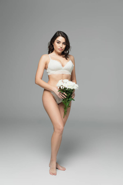 Pretty brunette woman in lace lingerie holding flowers and looking at camera on grey background  - Φωτογραφία, εικόνα