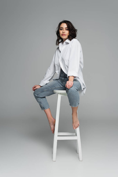 Pretty woman in jeans and shirt on chair on grey background  - Photo, Image