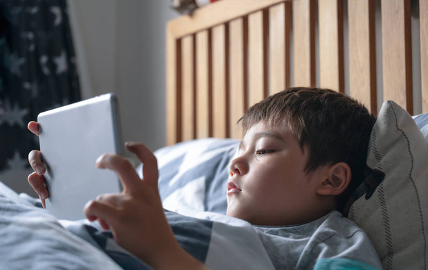 Niño leyendo cuentos de la hora de acostarse en la tableta antes de dormir, niño feliz sentado en la cama jugando juegos en la almohadilla digital, niño relajándose en casa en su dormitorio los fines de semana. - Foto, imagen