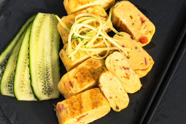 Traditional Japanese Tamagoyaki omelet. Pieces of tamago-yaki on a slate board with sliced cucumbers, close-up, top view - Photo, Image