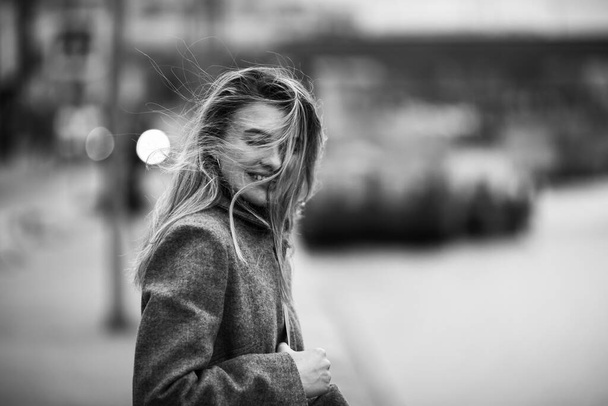Black and white city portrait of young happy, smiling attractive woman with long hair covering her face from strong wind - Photo, Image