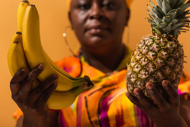 Bananen und Ananas in den Händen einer verschwommenen Afroamerikanerin mittleren Alters auf Orange - Foto, Bild