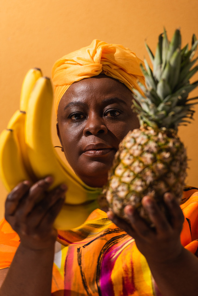 african american woman in yellow turban and colorful dress holding pineapple and bananas on orange - Φωτογραφία, εικόνα