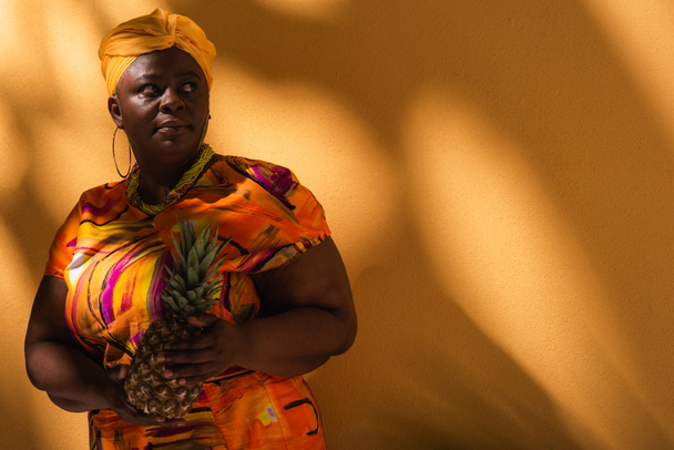middle aged african american woman in yellow turban and colorful dress holding pineapple on orange - Foto, Imagem