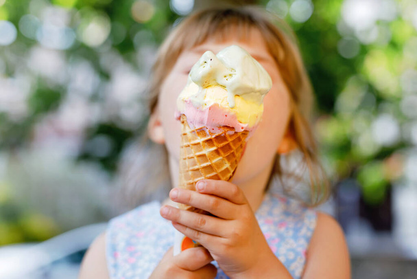 Little preschool girl eating ice cream in waffle cone on sunny summer day. Happy toddler child eat icecream dessert. Sweet food on hot warm summertime days. Bright light, colorful ice-cream - Foto, imagen