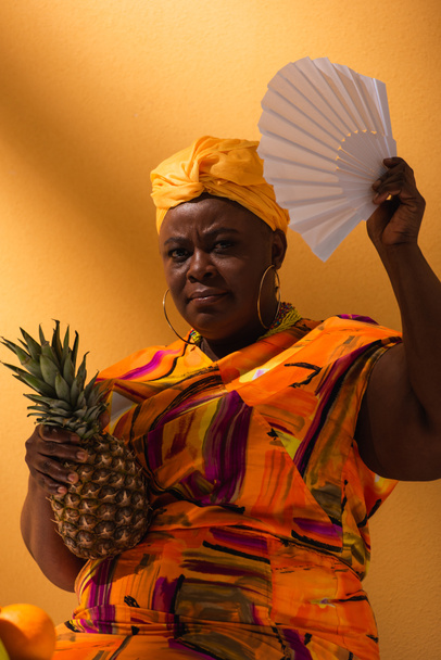 serious middle aged african american woman holding pineapple and waving with fan on orange - Photo, Image