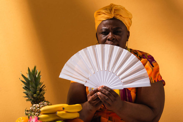 ernstige middelbare leeftijd Afrikaanse Amerikaanse vrouw zitten in de buurt van fruit en het houden van ventilator op oranje - Foto, afbeelding