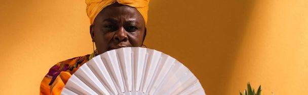 poważna Afroamerykanka w średnim wieku przykrywająca twarz wentylatorem na pomarańczowym, baner - Zdjęcie, obraz