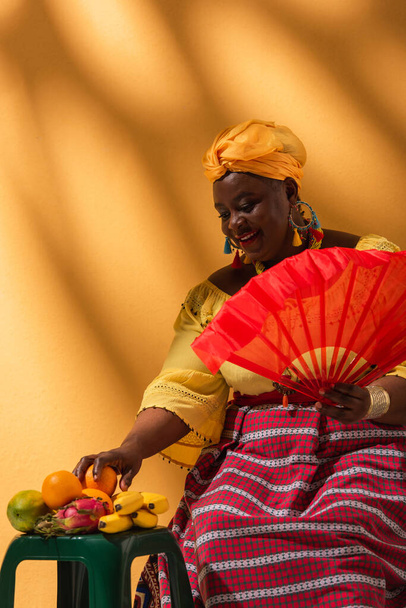 cheerful middle aged african american woman touching fruits and holding fan on yellow - Фото, изображение