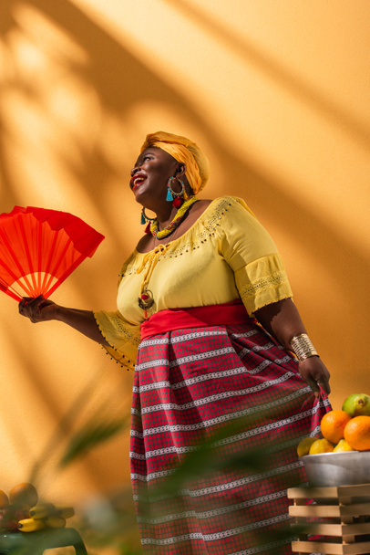 low angle view of positive middle aged african american woman selling fruits and holding fan on orange - Foto, immagini