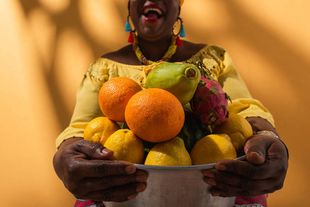 partial view of middle aged african american woman holding metal bowl with fruits on orange - Foto, Bild