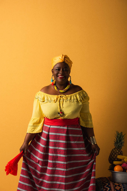 mulher afro-americana de meia idade em roupas brilhantes e turbante em pé perto de frutas em tigela de metal em amarelo - Foto, Imagem