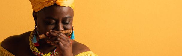 middle aged african american woman in yellow turban smelling aroma of cigar with closed eyes on yellow, banner - Foto, Imagem
