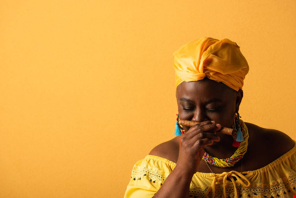 middle aged african american woman in yellow turban smelling aroma of cigar with closed eyes on yellow - Fotografie, Obrázek