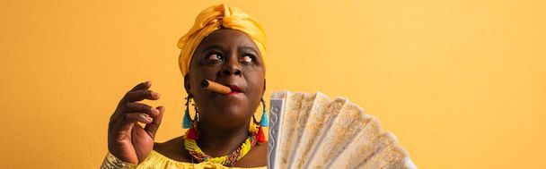 middle aged african american woman in yellow turban and blouse holding fan and smoking cigar on yellow, banner - 写真・画像
