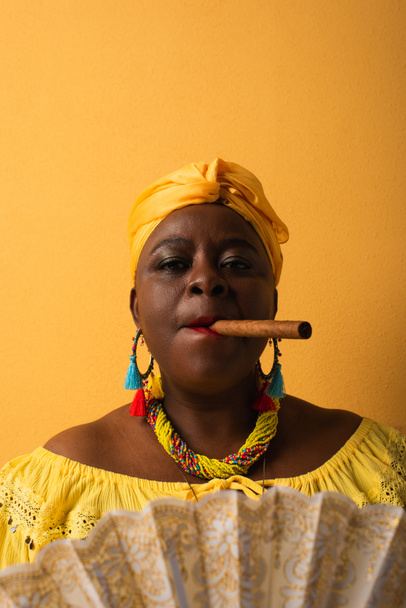 portret van ernstige middelbare leeftijd Afrikaanse Amerikaanse vrouw roken sigaar op geel - Foto, afbeelding
