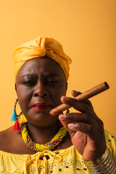 portrait of serious middle aged african american holding  cigar and looking at camera on yellow - Fotó, kép