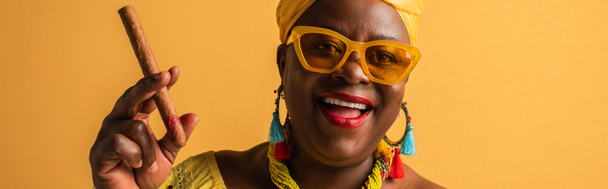 sorridente donna afroamericana di mezza età in occhiali da sole e accessori colorati che tengono il sigaro sul giallo, banner - Foto, immagini