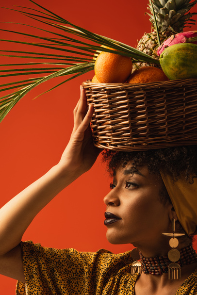 portrait of young african american woman holding basket with exotic fruits on head and looking away on red - Φωτογραφία, εικόνα