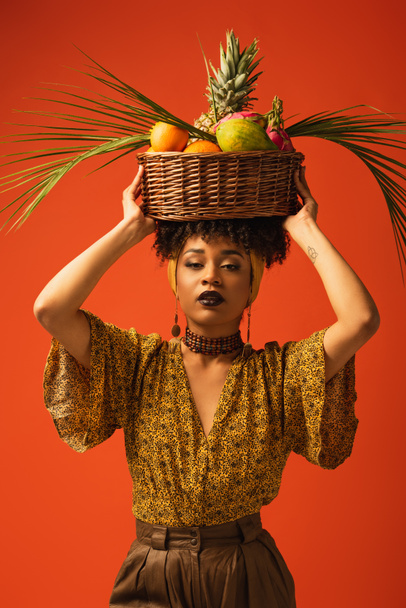 young african american woman holding basket with exotic fruits on head and looking at camera on orange - Fotografie, Obrázek