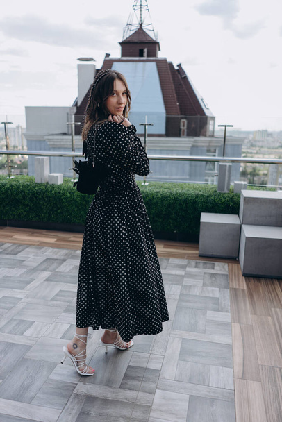 fashion photo of a beautiful woman dressed in a stylish black dress with white polka dots posing outdoor on a bright terrace - Фото, зображення