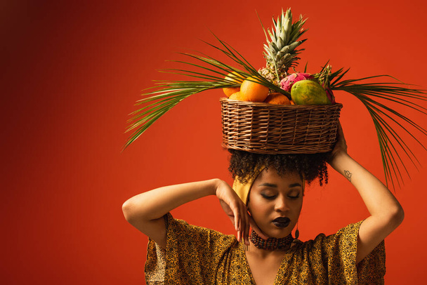 young african american woman with hand near face holding basket with exotic fruits on head on red - Fotografie, Obrázek