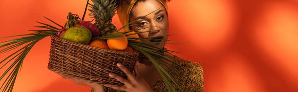 jong Afrikaans amerikaans vrouw houden mand met exotisch fruit in de buurt gezicht op oranje, banner - Foto, afbeelding