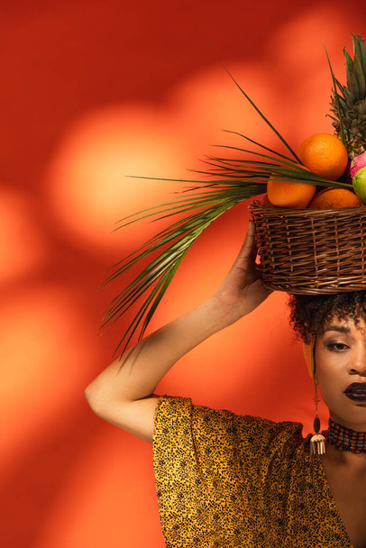 partial view of young african american woman holding basket with exotic fruits on head on orange - Zdjęcie, obraz
