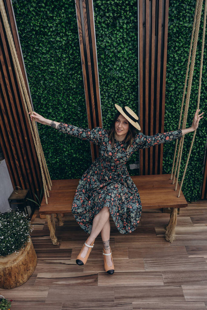 photo of a beautiful woman dressed in a stylish floral dress and straw hat posing on the wooden swing in the studio - 写真・画像