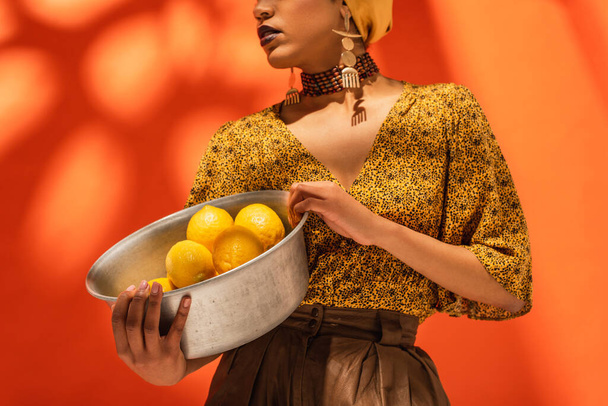 partial view of african american woman in yellow blouse holding metal bowl with lemons on orange - Photo, Image