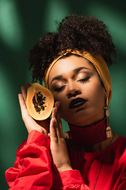 young african american woman with closed eyes holding half cut of ripe papaya near face on green - Фото, изображение