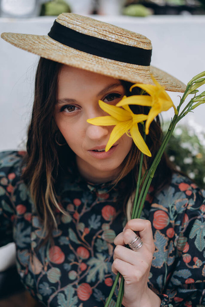 photo of a beautiful woman dressed in a stylish floral dress and straw hat posing with yellow flowers in the studio - Foto, immagini