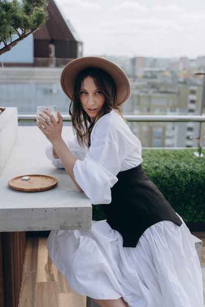 fashion photo of a cheeky woman dressed in a stylish white dress with a black designer belt posing with drink outdoor on a bright terrace - Photo, image