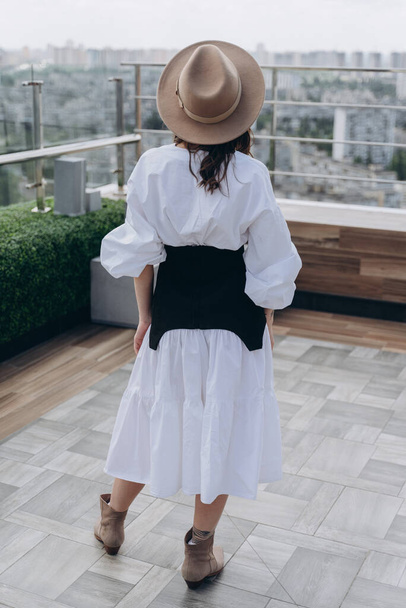 back view of a woman dressed in a stylish white dress with a black designer belt posing outdoor on a bright terrace - Foto, afbeelding