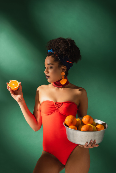 african american woman in red swimsuit holding cut orange and metal bowl with fruits on green - Foto, afbeelding