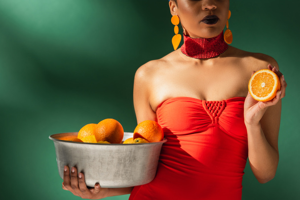 partial view of african american woman in red swimsuit holding cut orange and metal bowl with fruits on green - Foto, Imagen