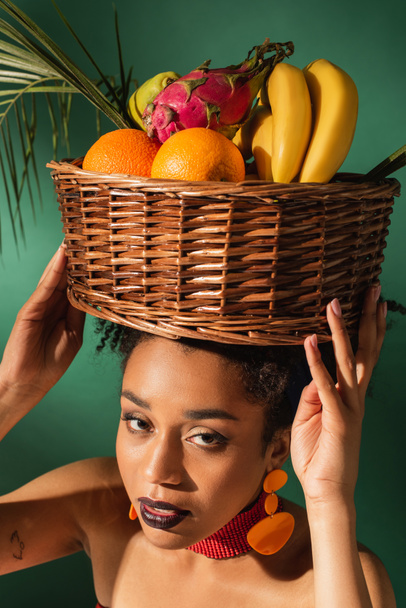 high angle view of pretty young african american woman holding basket with exotic fruits on head on green - Photo, Image