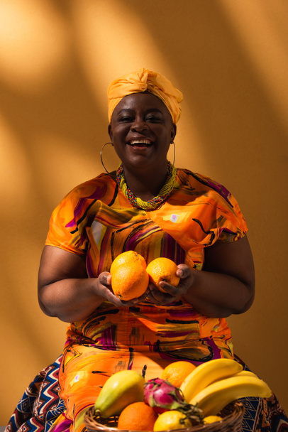 glimlachende middelbare leeftijd Afrikaanse verkoopster zittend met fruit op oranje - Foto, afbeelding