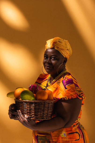 sonriente mujer afroamericana de mediana edad sosteniendo cesta con frutas exóticas en naranja - Foto, imagen