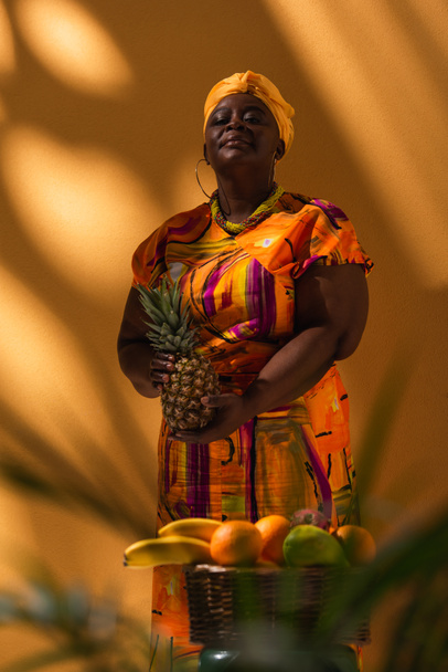 confident middle aged african american saleswoman standing near fruits with pineapple in hands on orange - Fotografie, Obrázek