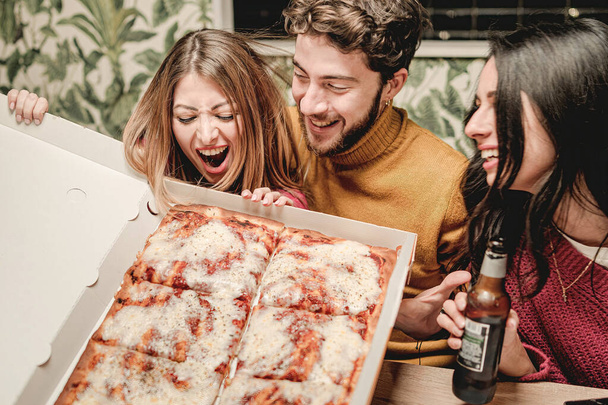 Groupe de jeunes gens s'amusant ensemble tenant une boîte de carton de pizza et plaisantant ensemble à la maison faisant une fête - Photo, image