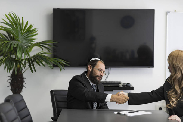 Dois parceiros de negócios israelitas a apertar as mãos no escritório. Homem judeu em um yarmulke e óculos sentados com mulher com cabelo loiro. Judeu de camisa, fato e chapéu nacional Kippah. Aperto de mão de duas mãos - Foto, Imagem